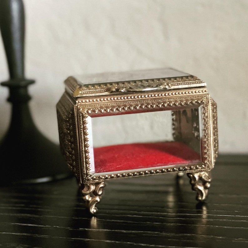 May include: A small, ornate, gold-colored jewelry box with clear glass sides and a red velvet interior. The box has decorative detailing and rests on four curved feet. The lid is hinged and has a small handle.