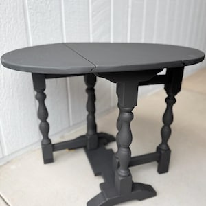 Puede incluir: Una mesa redonda gris oscuro con hojas abatibles y patas torneadas. La mesa está pintada con un acabado mate y tiene un diseño clásico y vintage. La mesa tiene aproximadamente 76 cm de diámetro cuando está completamente extendida.