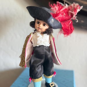 Könnte beinhalten: Eine Vintage-Puppe in einem Kostüm, mit einem schwarzen Hut und roter Feder. Die Puppe trägt ein weißes Hemd mit Spitze, eine schwarze Hose und einen goldfarbenen Umhang. Die Puppe steht auf einer blauen Schachtel.