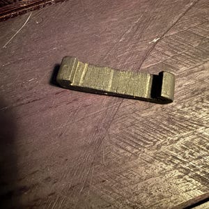 Op de afbeelding: Een donkergroen, rechthoekig object met een textuuroppervlak en een sleuf aan één uiteinde. Het object lijkt gemaakt te zijn van hout of een vergelijkbaar materiaal. Het object ligt op een donker, getextureerd oppervlak.