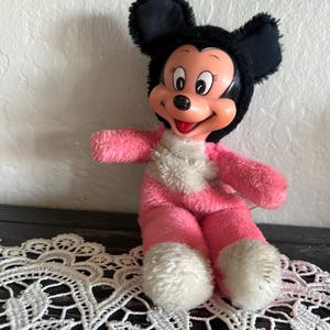 May include: A vintage Mickey Mouse plush toy. The toy has a black head with large ears, a pink body, and white accents. The plush is sitting on a white lace doily.