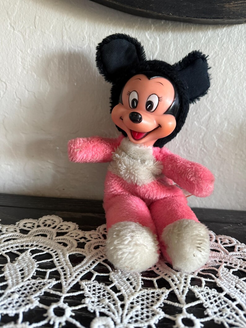 May include: A vintage Mickey Mouse plush toy. The toy has a black head with large ears, a pink body, and white accents. The plush is sitting on a white lace doily.