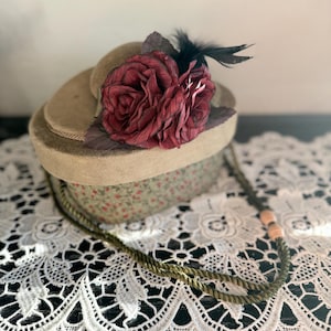 May include: A vintage-style hatbox with a floral pattern base and a tan lid. The hat is adorned with large burgundy fabric roses and black feathers. A decorative rope necklace with peach-colored beads rests in front of the hatbox.