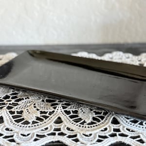 May include: A rectangular, glossy black ceramic serving tray. The tray has rounded corners and a slightly raised edge. It sits on a white lace doily, with a blurred white background.