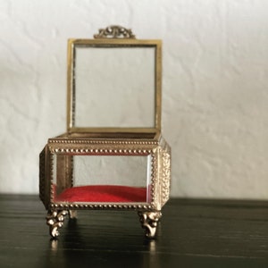 May include: A small, open antique jewelry box with a gold-colored metal frame and clear glass panels. The interior features a vibrant red velvet cushion. The box rests on ornate, curved feet and has a decorative top.