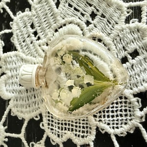 May include: A clear, heart-shaped glass bottle with a white stopper, filled with white flowers and green leaves. The bottle is set on a white lace doily, creating a delicate and natural aesthetic. The bottle's contents are visible through the transparent glass.