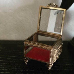 May include: An open, antique-style jewelry box with a gold-colored metal frame and clear glass panels. The interior is lined with red fabric. The box is resting on four ornate feet and has decorative detailing.