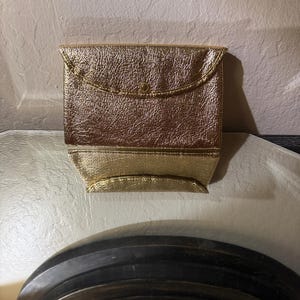 Op de afbeelding: Een goudkleurige, getextureerde clutch met een klepsluiting en een kleine knoop. De clutch heeft een rechthoekige vorm met een gevouwen bodem. Het materiaal lijkt metaal of imitatieleer te zijn en reflecteert licht.