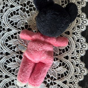 May include: A plush toy in the shape of a mouse, featuring a black head and pink body. The toy has white accents on the feet and is made of a soft, fuzzy material. The toy is lying on a white lace doily.