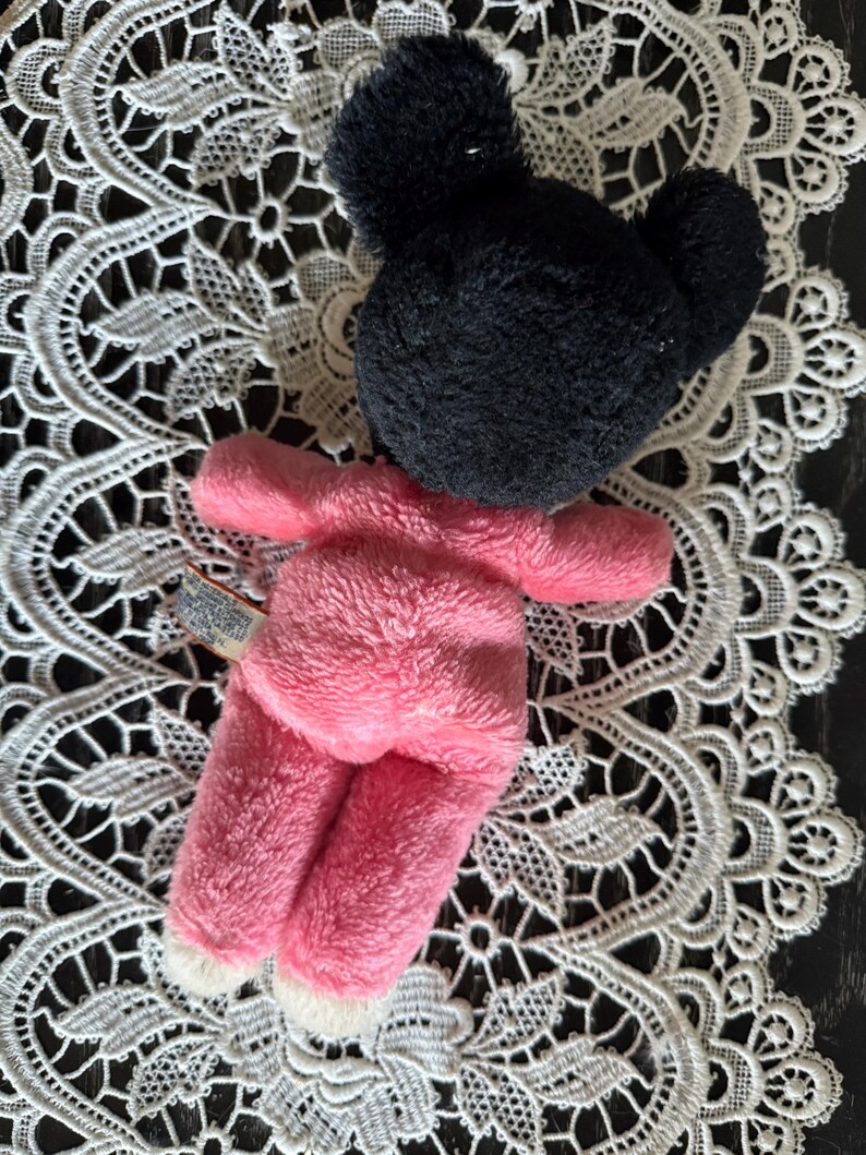 May include: A plush toy in the shape of a mouse, featuring a black head and pink body. The toy has white accents on the feet and is made of a soft, fuzzy material. The toy is lying on a white lace doily.