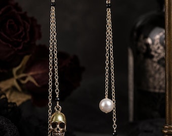 May include: A pair of gold-toned drop earrings featuring a hook closure, a clear round crystal, and two delicate chains. One chain ends in a metallic skull charm, while the other features a white pearl bead, accented with small black beads.