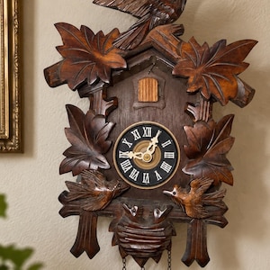 May include: A dark brown wooden cuckoo clock featuring carved maple leaves, a bird perched at the top, two birds on the sides, and a nest with two small birds at the bottom. The clock face has a black background with white Roman numerals.
