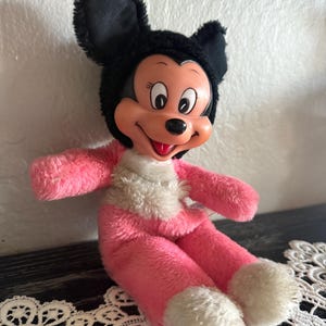 May include: A vintage Minnie Mouse plush doll. The doll has a black head and ears, a pink body, and white accents on the feet and chest. The doll has a smiling face with black eyes and a red mouth.