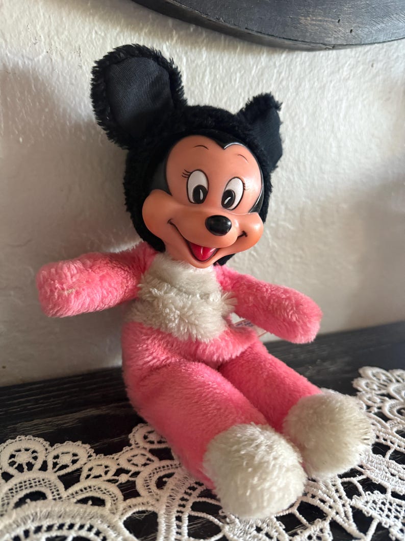 May include: A vintage Minnie Mouse plush doll. The doll has a black head and ears, a pink body, and white accents on the feet and chest. The doll has a smiling face with black eyes and a red mouth.