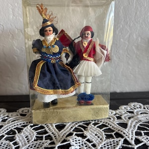 May include: Two vintage Greek dolls in traditional attire, displayed in a clear plastic box. The dolls are dressed in detailed costumes with gold accents, including hats, dresses, and vests. One doll wears a navy blue dress, and the other wears a red vest and white skirt.