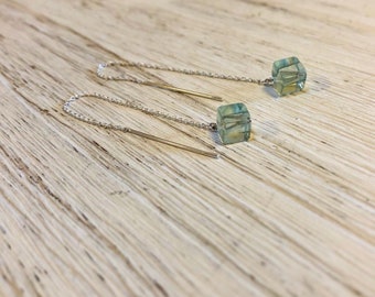 May include: A pair of delicate earrings featuring a small, translucent, cube-shaped bead in shades of green and blue. The bead is suspended from a fine silver chain and a gold-colored post. The earrings are displayed on a light-colored wooden surface.