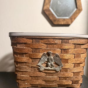 May include: A brown woven basket with a metal eagle emblem, set on small wooden feet. The basket has a rectangular shape and a dark rim. An octagonal mirror with a wooden frame hangs in the background.