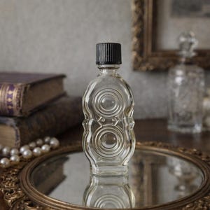 May include: A clear glass perfume bottle with a black cap, featuring a decorative design of concentric circles. The bottle sits on a gold-rimmed mirror, with antique books and pearls in the background.