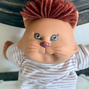 Peut inclure: Poupée vintage avec un visage de chat, peau brune et cheveux en fil rouge. Elle porte une chemise rayée et un pantalon en jean bleu. Les bras de la poupée sont tendus, et elle a des traits peints, évoquant la nostalgie.