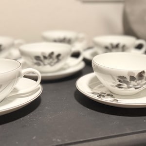 Puede incluir: Juego de tazas y platillos blancos. Cada taza y platillo presenta un diseño floral negro. Las tazas tienen asa y se apoyan en un platillo a juego. El juego incluye seis tazas y platillos, perfecto para el té.