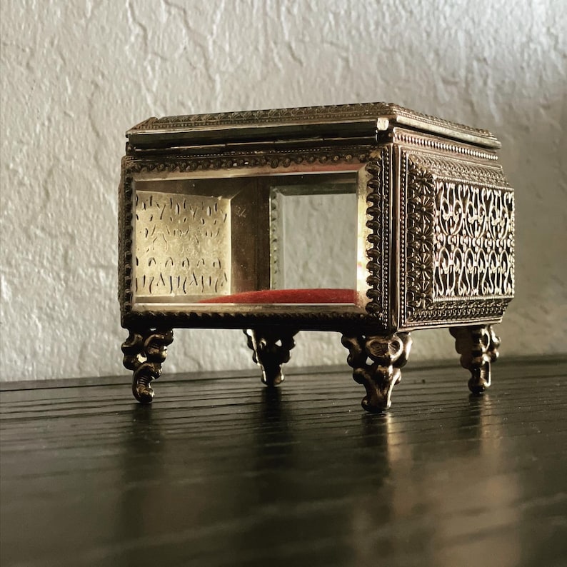 May include: A small, antique display box with a rectangular glass enclosure and ornate metal filigree detailing. The box has a red velvet interior and rests on four decorative, curved legs. The metal has a tarnished, aged appearance.
