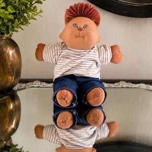 Op de afbeelding: Een pluche kattenpop met een gezicht, oren en pootjes van beige stof, voorzien van een pluk roodbruin garen als haar. De pop draagt een wit en beige gestreept shirt met korte mouwen en een donkerblauwe spijkerbroek, met een reflectie eronder.