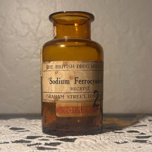 Könnte beinhalten: Eine antike Apothekerflasche aus braunem Glas mit einem Etikett mit der Aufschrift "Sodium Ferrocyanide" und "Poison". Das Etikett enthält auch "The British Drug Houses" und "Graham Street, London". Die Flasche hat einen kleinen, runden Hals und einen leicht ausgestellten Rand.