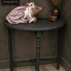 May include: A semi-circular dark gray console table with four turned legs and a lower support structure. On the table sits a plush animal figure wearing a lavender floral dress with a white lace collar and a matching lavender ribbon bow.