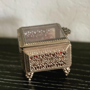 May include: A small, ornate silver-colored jewelry box with a glass top. The box features intricate filigree detailing on the sides and a red velvet interior. It rests on four small, decorative feet.