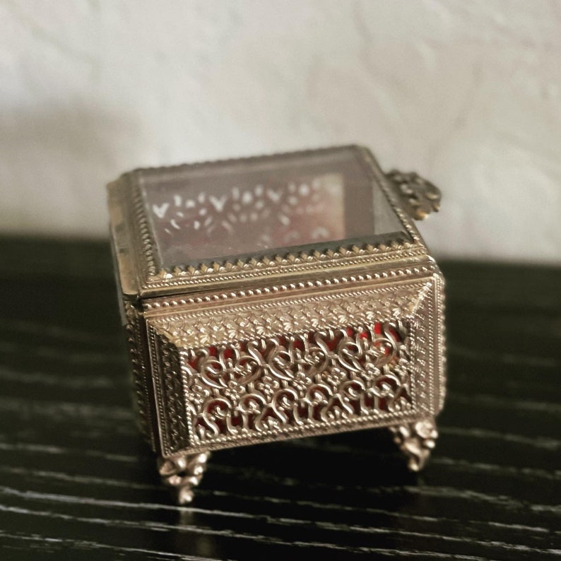 May include: A small, ornate silver-colored jewelry box with a glass top. The box features intricate filigree detailing on the sides and a red velvet interior. It rests on four small, decorative feet.