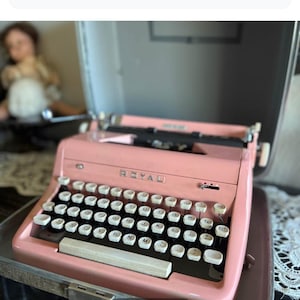 Può includere: Una macchina da scrivere Royal Quiet Deluxe rosa. La tastiera nera ha tasti bianchi. La parola "ROYAL" è visibile. La macchina da scrivere è in una custodia argentata. Un oggetto vintage per collezionisti.