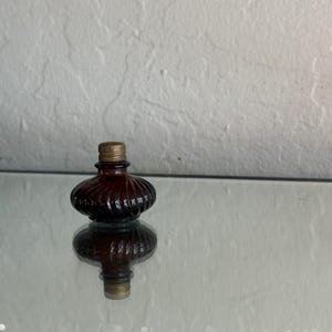 May include: A small, antique-style brown glass bottle with a ribbed design and a gold-colored metal cap. The bottle is sitting on a reflective surface, creating a mirrored effect. The bottle's shape is round with a flat base.