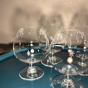 Puede incluir: Un conjunto de copas de brandy transparentes con diseños de estrellas grabadas. Las copas tienen forma redondeada, un tallo corto y una base circular. Se disponen sobre una bandeja de color verde azulado, reflejando la luz y creando una sensación elegante.