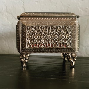 May include: A small, ornate silver-colored jewelry box with intricate floral filigree detailing. The box has a hinged lid and rests on four decorative feet. The interior appears to be lined with red fabric.