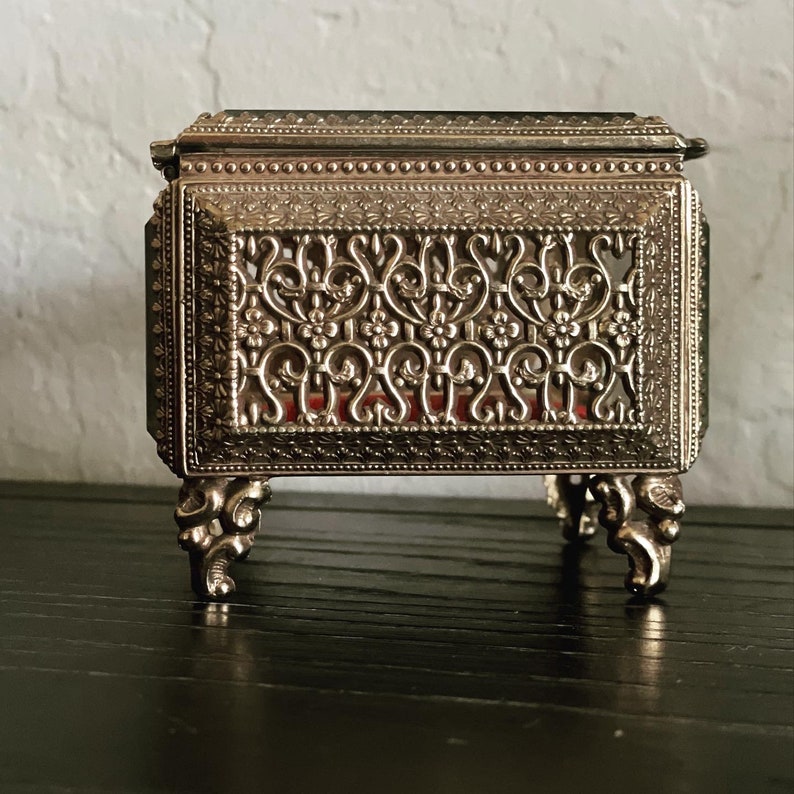 May include: A small, ornate silver-colored jewelry box with intricate floral filigree detailing. The box has a hinged lid and rests on four decorative feet. The interior appears to be lined with red fabric.