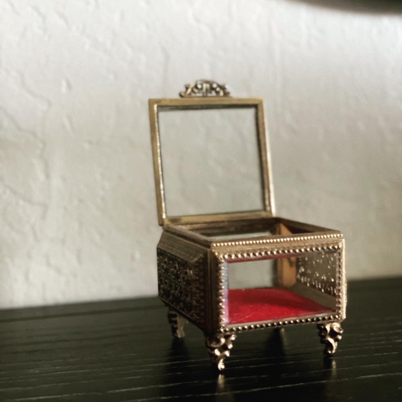 May include: An open, antique-style jewelry box with a hinged glass lid. The box is gold-toned with ornate detailing and rests on four decorative feet. The interior is lined with red velvet.