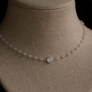 May include: A delicate necklace with small, round, pale gemstones strung on a fine chain. A larger, round, iridescent stone is centered on the necklace. The necklace is displayed on a neutral-toned mannequin.