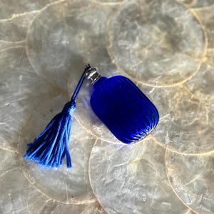 May include: A cobalt blue glass perfume bottle with a ribbed design and a matching blue tassel. The bottle has a silver-colored metal cap. The tassel is attached to the bottle with a thin blue cord.