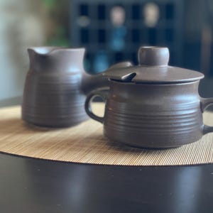 Può includere: Un set di lattiera e zuccheriera con coperchio in ceramica marrone. La lattiera ha un beccuccio e un manico. Lo zuccheriera ha due manici e una fessura per il cucchiaio nel coperchio. Entrambi i pezzi presentano dettagli orizzontali a coste.