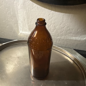Könnte beinhalten: Eine Vintage-Braunglasflasche mit dem geprägten Wort "CLOROX" wird auf einem silbernen Tablett präsentiert. Die Flasche hat eine abgerundete Schulter und einen schmalen Hals. Die Flasche ist wahrscheinlich antik und zeigt ein klassisches Design.