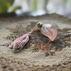 May include: A vintage perfume bottle with a pink, petal-shaped design and a matching tassel. The bottle has a gold-colored cap and is set on a decorative, embroidered doily with a gold trim. The overall aesthetic is delicate and antique.