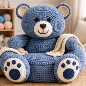 May include: A blue crocheted bear-shaped chair featuring a large head with black eyes, a cream-colored muzzle, and a brown nose. The chair has two front-facing feet with cream and blue paw pads and a cream-colored fabric draped over the arms.