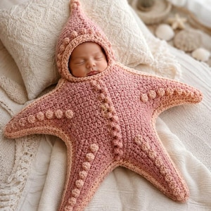 May include: A crocheted baby swaddle blanket shaped like a five-pointed starfish in a dusty rose color. The garment features a pointed hood, a central vertical seam, and raised bobble stitch textures along the edges and center of each arm.