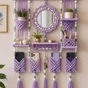 Peut inclure: Un organisateur mural en macramé de couleur lavande avec un miroir circulaire central, plusieurs étagères et quatre poches contenant des smartphones. L'ensemble est décoré de perles blanches et vertes, de pompons et divers petits objets.