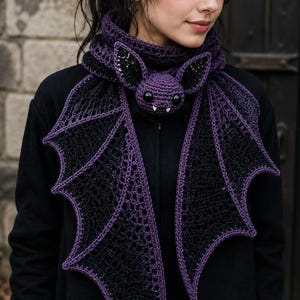 May include: A crocheted scarf featuring a central purple bat head with black and white eyes and two small white fangs. The scarf extends into two large, black, wing-shaped panels with purple crocheted edges, draped around the neck area.