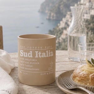 Puede incluir: Una taza de cerámica beige con texto blanco que dice THE COFFEE EDIT, Sud Italia, caffè napoletano, caffè alla nocciola, caffè leccese, caffè ammantecato, No. 1414 y EDIT & STONE, junto con un pequeño dibujo lineal de una cafetera.