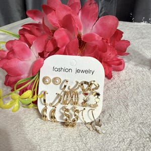 Op de afbeelding: Een set goudkleurige oorbellen op een witte displaykaart met de tekst fashion jewelry. De collectie bevat parelknopjes, gladde ringen, ringen met textuur, gedraaide metalen ontwerpen en kleine bungelende vlinderbedels in diverse vormen.
