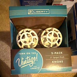 May include: A two-pack of Liberty brand 2-inch spigot knobs in a teal and brown cardboard box. The knobs are white, round, and feature a decorative, open-work metal design resembling a vintage valve handle. The box text reads Turn on the Vintage! with LIBERTY and 2-PACK 2" Spigot Knob KNOBS.