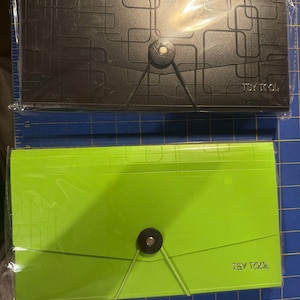 May include: Two rectangular document organizers, one black and one lime green, each featuring a geometric square pattern, a central circular button closure with an elastic cord, and the text TSY TOOL printed in the bottom right corner.