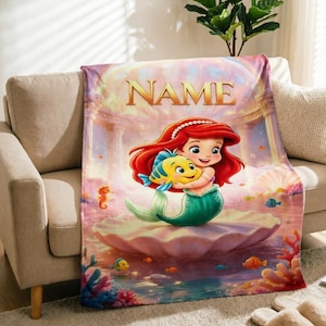 Puede incluir: Una manta suave con una sirena de dibujos animados de pelo rojo largo y cola verde, que sostiene un pez amarillo y azul dentro de una concha grande. La palabra NAME está impresa en oro en la parte superior, rodeada de coral y vida marina.